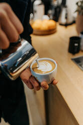 Ein anonymer Barista gießt Milch in eine Tasse Kaffee, während er in der Cafeteria einen Milchkaffee zubereitet - ADSF24660