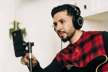 Stativ mit Kamera in der Nähe von Mann spielt Gitarre im Haus Zimmer platziert - ADSF24674