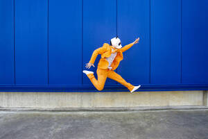 Mann in leuchtend orangefarbenem Anzug und Pandamaske springt gegen eine blaue Wand - OIPF00697