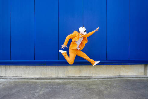 Man wearing vibrant orange suit and panda mask jumping against blue wall - OIPF00697