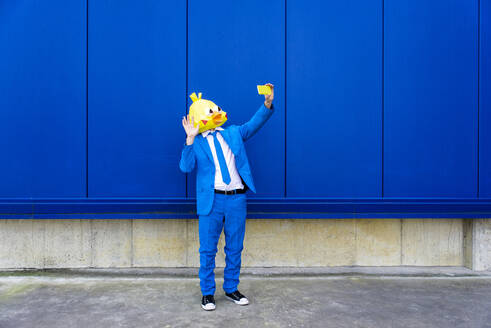 Mann in leuchtend blauem Anzug und Vogelmaske macht ein Selfie mit seinem Smartphone vor einer blauen Wand - OIPF00723