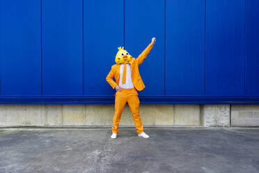 Mann in leuchtend orangefarbenem Anzug und Vogelmaske steht vor einer blauen Wand mit erhobener Hand - OIPF00730