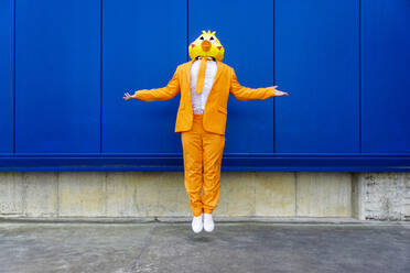 Mann in leuchtend orangefarbenem Anzug und Vogelmaske schwebend vor einer blauen Wand - OIPF00732