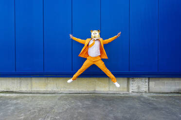 Mann in leuchtend orangefarbenem Anzug und Nagetiermaske springt gegen eine blaue Wand - OIPF00734