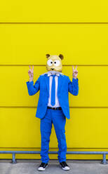 Mann in leuchtend blauem Anzug und mit Nagetiermaske, der vor einer gelben Wand ein Friedenszeichen macht - OIPF00737