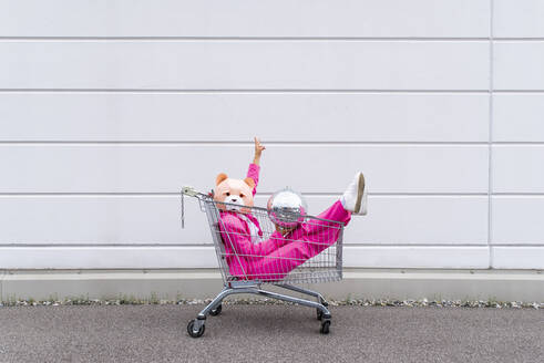 Frau in pinkfarbenem Anzug und Bärenmaske sitzt in einem Einkaufswagen mit Discokugel in den Händen - OIPF00809