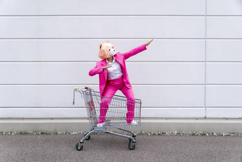 Frau in leuchtend rosa Anzug und Bärenmaske steht mit erhobenen Armen im Einkaufswagen - OIPF00810