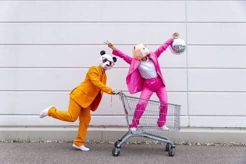 Mann und Frau in farbenfrohen Anzügen und Tiermasken, die mit Einkaufswagen und Discokugel herumspielen - OIPF00813