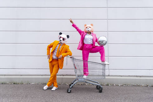 Mann und Frau in farbenfrohen Anzügen und Tiermasken posieren mit Einkaufswagen und Discokugel vor einer weißen Wand - OIPF00815