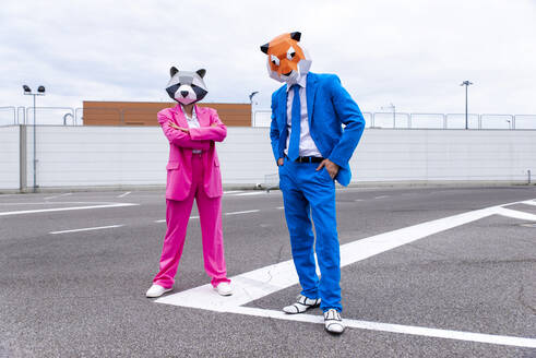 Mann und Frau in farbenfrohen Anzügen und Tiermasken posieren Seite an Seite auf einem leeren Parkplatz - OIPF00817