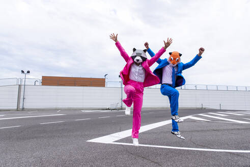 Mann und Frau in farbenfrohen Anzügen und Tiermasken springen nebeneinander auf einem leeren Parkplatz - OIPF00818