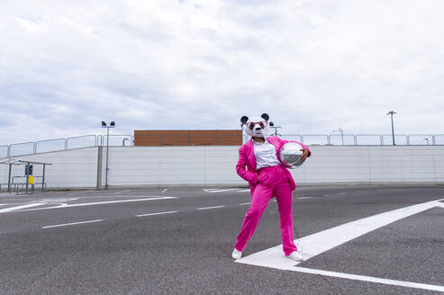 Frau in leuchtend rosa Anzug und Panda-Maske posiert auf leerem Parkplatz mit Discokugel in der Hand - OIPF00820