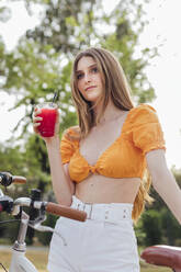 Thoughtful woman holding glass of juice by bicycle - JRVF00784