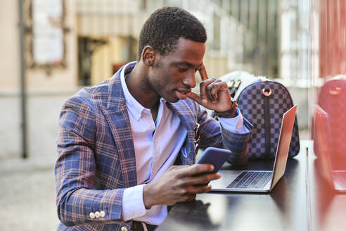 Junger männlicher Unternehmer mit Mobiltelefon und Laptop in einem Straßencafé - AODF00494