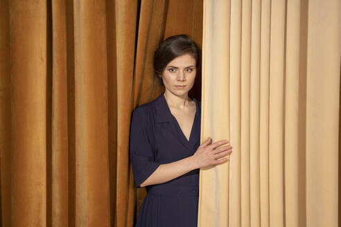 Confident young woman standing by curtain - VPIF04051