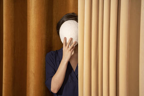 Woman covering face with white mask by curtain - VPIF04052