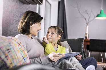 Smiling mother with digital tablet looking at daughter while sitting on sofa - AUF00607