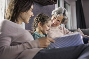 Daughter looking at photo album while sitting by family in living room - AUF00615