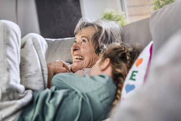 Cheerful grandmother lying by granddaughter on sofa at home - AUF00620