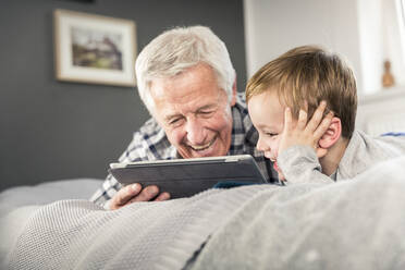 Lächelnder Großvater und Enkel benutzen ein digitales Tablet, während sie auf dem Bett liegen - AUF00655