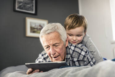 Grandson using digital tablet with grandfather on bed at home - AUF00656