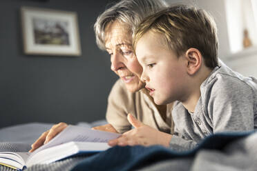 Grandmother reading book while lying by grandson in bedroom - AUF00658