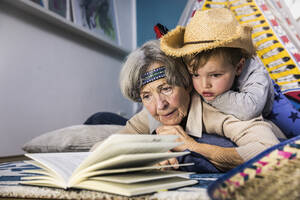 Senior woman reading book while boy lying on her at home - AUF00662
