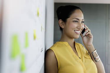 Lächelnde Geschäftsfrau, die wegschaut, während sie mit dem Handy im Büro telefoniert - BMOF00741