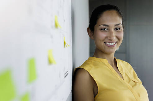 Lächelnde Geschäftsfrau, die sich im Büro an die Wand lehnt - BMOF00742