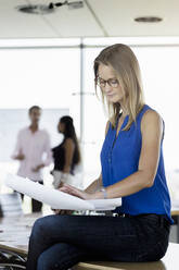 Businesswoman working on blueprint while sitting on desk with male and female colleagues discussing in background at office - BMOF00745