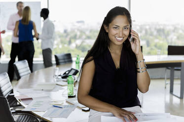 Lächelnde Geschäftsfrau im Gespräch am Handy mit männlichen und weiblichen Kollegen, die im Hintergrund im Büro diskutieren - BMOF00747