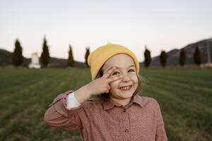 Smiling girl wearing knit hat gesturing peace sign while looking away - RCPF01099