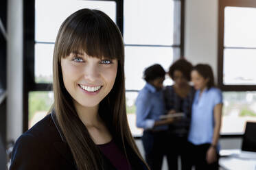 Smiling beautiful businesswoman with bangs standing while male and female colleagues discussing in office - BMOF00785