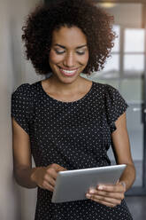Lächelnde, lockig behaarte Unternehmerin, die im Büro stehend ein digitales Tablet benutzt - BMOF00786