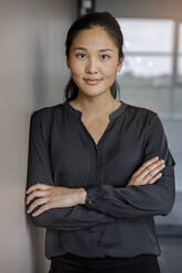 Smiling young confident businesswoman standing with arms crossed in office - BMOF00788