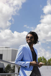 Lächelnder junger Geschäftsmann mit Smartphone, der an einem sonnigen Tag am Geländer steht und wegschaut - BMOF00796