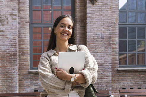 Lächelnde Frau mit Laptop vor einem Haus stehend - PNAF01655