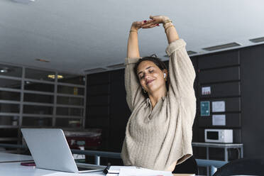 Lächelnde weibliche Fachkraft mit geschlossenen Augen, die ihre Hände ausstreckt, während sie im Büro vor einem Laptop sitzt - PNAF01663