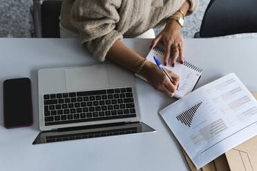 Businesswoman writing on diary while working on financial data at office - PNAF01666