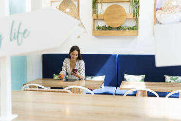 Smiling woman using smart phone while having juice in restaurant - PNAF01672