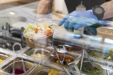 Koch mit Schutzhandschuhen bei der Zubereitung von Salat an der Essensausgabe im Restaurant - PNAF01681