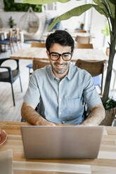 Smiling young businessman using laptop at table in cafe - RCPF01110