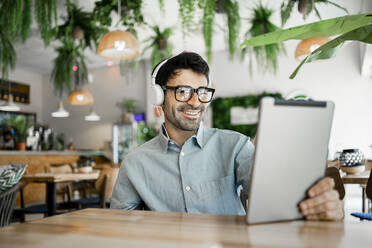 Lächelnder Geschäftsmann mit Kopfhörern, der in einem Restaurant ein digitales Tablet benutzt - RCPF01112