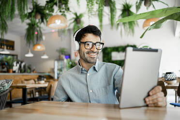 Smiling businessman wearing headphones using digital tablet at restaurant - RCPF01112