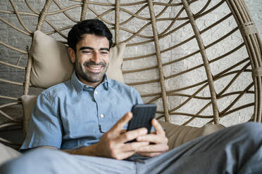 Lächelnder Geschäftsmann, der ein Mobiltelefon benutzt, während er auf einer Bambusschaukel sitzt - RCPF01116