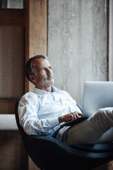 Senior male entrepreneur with laptop looking away while contemplating in office - GUSF05770