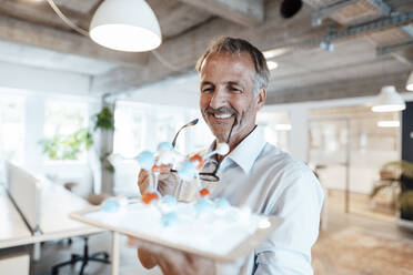 Smiling businessman holding digital tablet and artificial molecule in office - GUSF05775