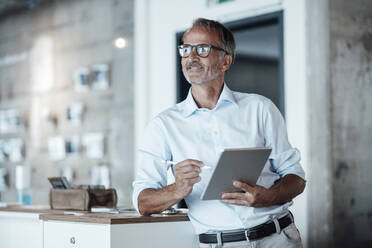 Thoughtful businessman with digital tablet looking away while standing in office - GUSF05777