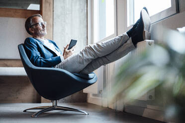 Geschäftsmann, der ein Mobiltelefon hält und auf einem Stuhl im Büro sitzt - GUSF05785