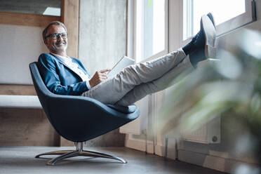 Männlicher Unternehmer mit digitalem Tablet entspannt auf einem Stuhl im Büro - GUSF05787